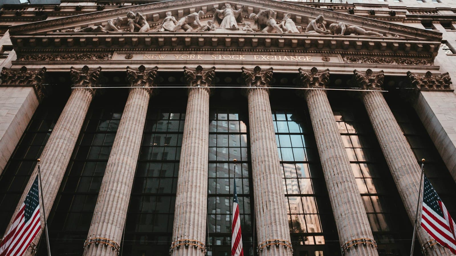 The facade of the New York Stock Exchange seen from below, its columns behind three American flags