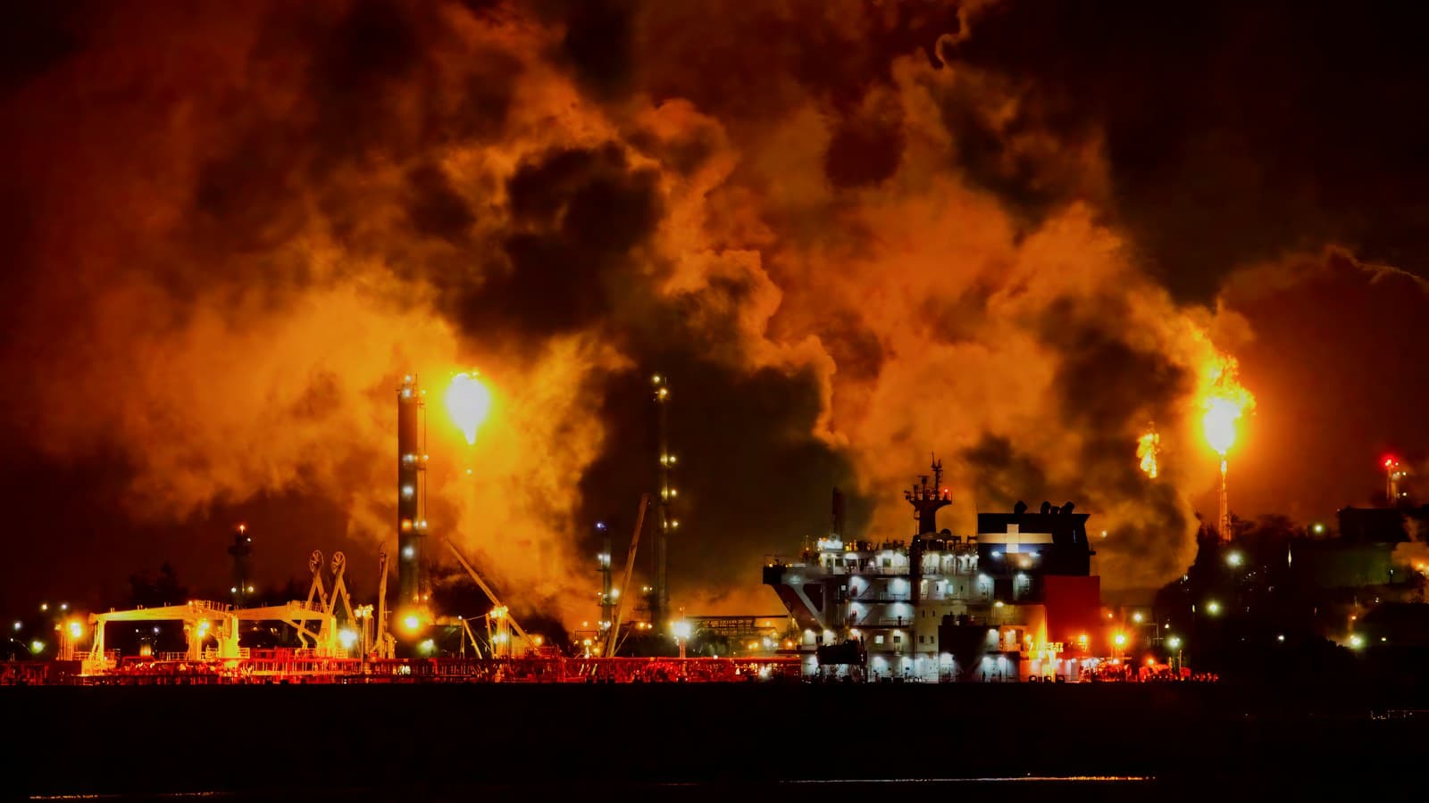 An oil refinery at night, its flares lighting the smoke orange over a tanker at the quay